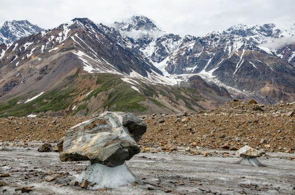 A landscape view of the Black Rapids Glacier in the Alaska Range, featuring large boulders balanced on narrow ice pedestals in a rocky glacial field. In the background, rugged mountains partially covered with snow and shrouded by clouds rise dramatically. The foreground shows a prominent boulder standing on a slender column of melting ice, with scattered stones and diminishing ice around it.