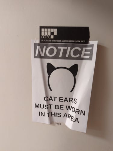 Piece of paper stuck to the wall. The paper says "Cat ears must be worn in this area". The tape used is CCCAC-branded.