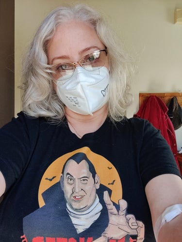 Me wearing Jeff Brawn's Count Floyd shirt, a character from SCTV played by Joe Flaherty. I have shoulder length ash blonde hair, gold glasses, and an N95 on with "T❤️" on it to one side. I have a bandage on my arm from a blood draw.