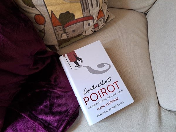 The book Poirot by Mark Aldridge over an armchair with a purple blanket and pillow with houses embroidered