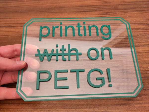Eine Durchsichtige Platte aus PETG, gehalten in einer Hand. Auf der Platte ist ein Text 3D gedruckt: "printing with on PETG!". Das "with" ist durchgestrichen.
