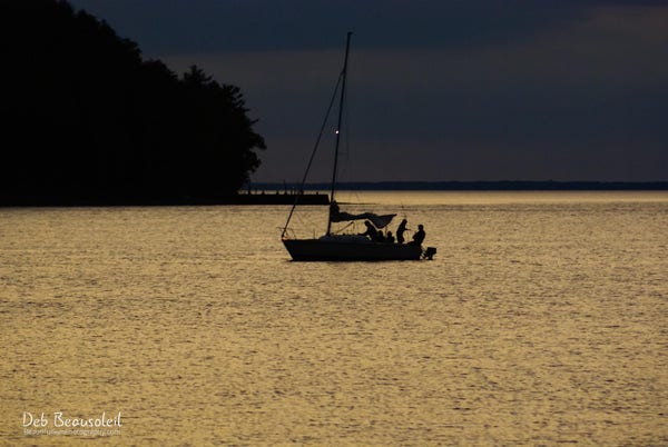 Photograph of a sailboat in Door County, Wisconsin, with figures on board visible against the gold-tinged waters of the bay at dusk, bordered by a silhouette of trees.  Image at:  https://beautifulsunphotography.com/featured/furling-the-sail-deb-beausoleil.html See more art & blog at: https://beautifulsunphotography.com/ https://debbeautifulsunphotography.com/ https://www.zazzle.com/store/beautifulsun_designs https://debbeausoleil.com