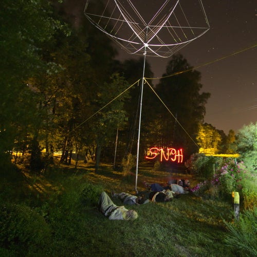 Night long exposition photo of contest team laying on the ground next to the hexbeam antenna on a mast, above them there is SN9H text painted by a person with red color headlight