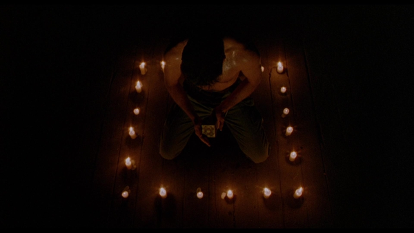 In a square of lit candles, a white man with short dark hair works to solve a puzzle box. He is bare chested and covered in sweat. The scene is otherwise dark.