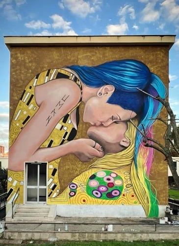 A large mural on a building depicting two people with stylized features sharing a kiss, with one person having blue hair and the other having blond hair with a colorful streak. The background is a blue sky with clouds.