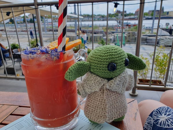 Crocheted Grogu doll stands adjacent to a glass of red / orange liquid (a beetroot smoothie) topped with blue flowers and orange slices, in the background the River Oder is visible, the sky is a mix of blue and white with scattered clouds