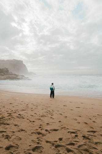 A beach with lots of mist, a man stands in the middle. In the background is an overcast sky and cliffs.