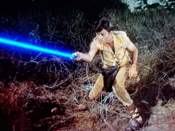 TOS scene. Bones is on planet somewhere. Foresty looking. He's wearing an all leather getup with a leather satchel. Here's, he's crouched and holding a lightsaber. JK fam he's firing a phaser but see it's Star Wars Day and it looks like a lightsab--you know what just forget it. 