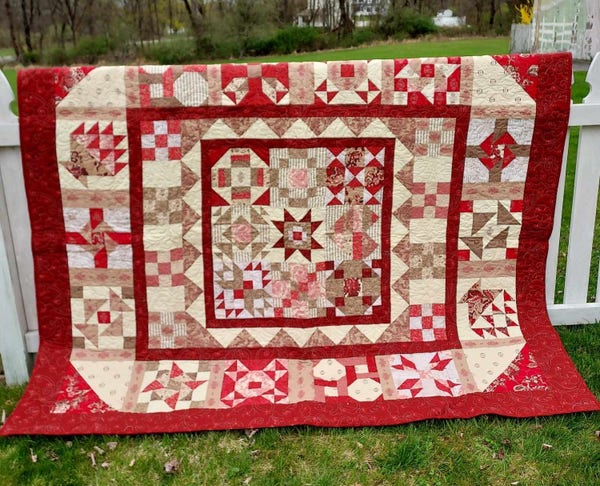 a photo of a quilt, it is different folksy geometric designs , rose-red on light tan, some checkered boxes with tan and white cubes in them, lots of pink and rosey patterns 
