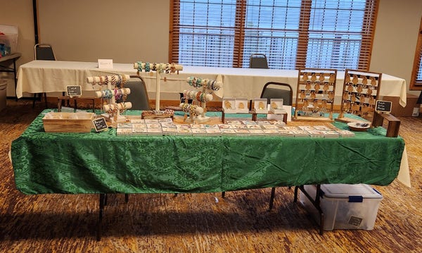 an eight foot long table with 2 racks of earrings for sale, 40 gift boxes displaying bracelets, and three risers with arms that display more bracelets