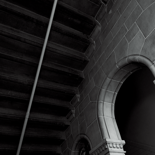 Black and white photo of the underside of a dark stone staircase on the left and a small neo-gothic archway on the right of the image.