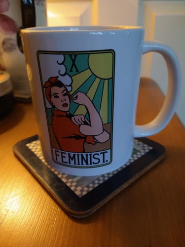 Mug of tea on a bedside cabinet, with an image of Rosie the riveter in a red shirt with matching bandana the word Feminist in black text at the bottom, a green and yellow sun top right and white fluffy cloud top left 