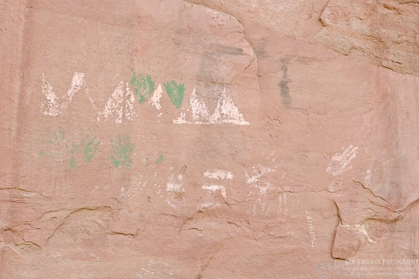 A color photo of a white and green pictograph on a dark tan sandstone cliff face. The white part is in two parallel lines. The top line begins on the left with a zigzag shaped that then four triangles. Two green upside down triangles fill in the space between the white triangles one, two, and three. Below the top line are what appear to be green hand prints, then some white markings, and then several white hand prints.