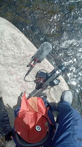 POV view of a man's leg and foot on a large boulder. The foot is next to a red/blue backpack, camera, and microphone. Surrounding the boulder is fast-flowing river water.