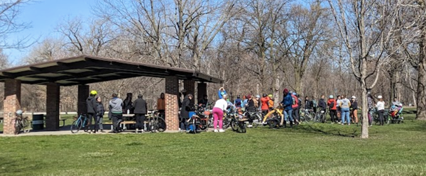 a large group of people with their bicycles in a park, near a picnic grove and some trees