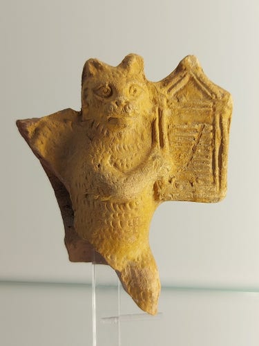 The picture shows a terracotta figure of a standing cat playing a harp with its front legs. The lower part of the figurine is broken off.
