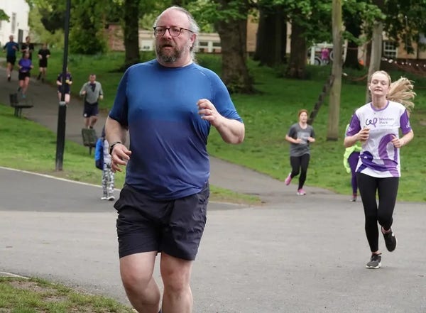 A man with a beard and grey hair running in a city park.