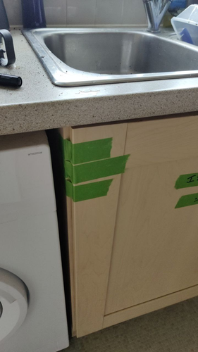 A cupboard door under a sink. It's broken on the top hinge and has been held together by green tape. It is clearly not a fantastic DIY effort.