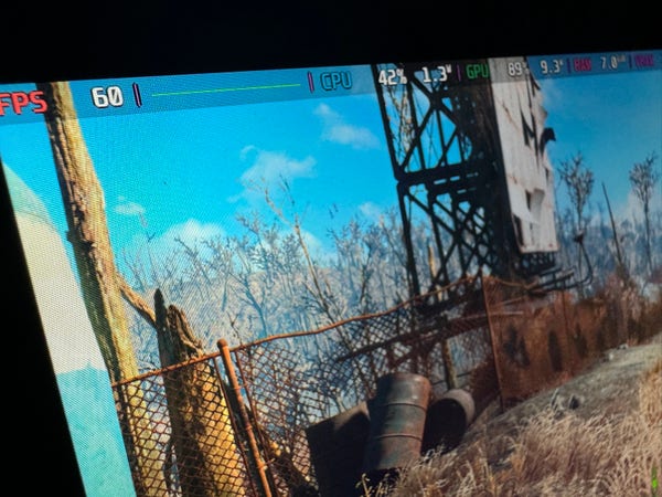 A SteamDeck screen displaying a video game scene with a watchtower, barrel, and a fence in a barren landscape. Performance metrics are visible at the top of the screen.