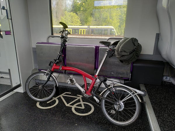 A red and black Brompton, unfolded in a bike space on a train. A green dry bag is bungee'd to the underside of the saddle. 