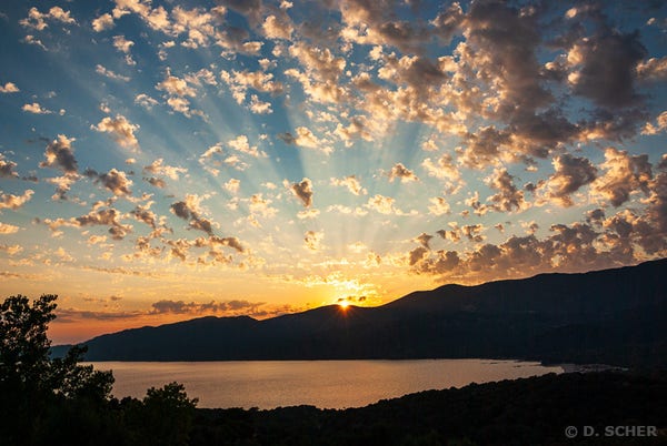 Sunset over a Corsican bay: the land and hill surrounding the bay are in shadow, and the calm waters of the bay have orange reflections.
The sun is setting behind a hill in the distance, sending bright rays into the sky dotted with small clouds casting their shadows.