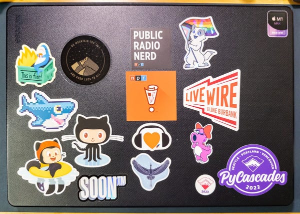 16-inch M1 Max MacBook Pro with a hard case decorated with stickers from NPR, GitHub, Live Wire Radio and PyCascades, along with stickers of Blahaj, a cat waving a LGBTQ+ pride flag, Birdo from Super Mario Bros. 2 (USA), SOON TM, and TBTL
