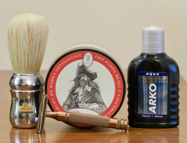A boar brush with a long loft and a chromed handle that bears a small Omega label stands next to a tub of shaving soap whose label shows a line drawing of a 17th-century pirate — with long messy hair, a long beard, a tricorn hat, and holding a long-stemmed clay pipe. a bronze-colored DE razor lying on its side in front obscrures the name of the soap ("Pirata"). To the right is a small blue-plastic bottle labeled "Arko Aqua."