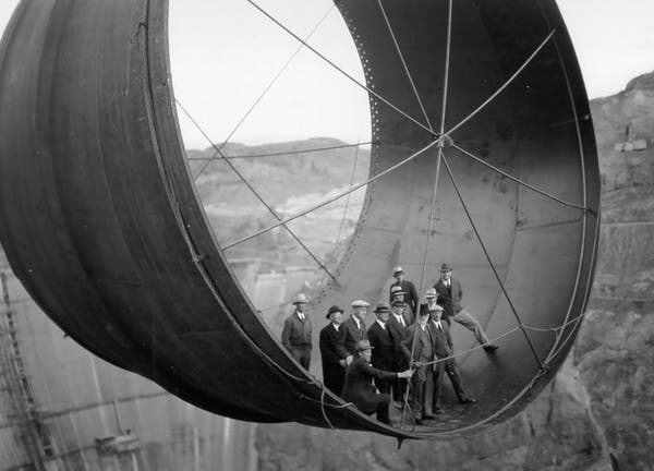 A group of men gathered in the lower end of a giant conduit, during the building of the Hoover Dam. The conduit is suspended high in the air.