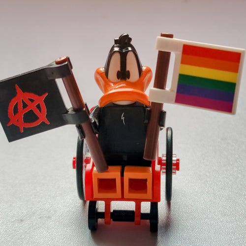 Une figurine Lego représentant Daffy Duck, dans un fauteuil roulant, avec un drapeau dans chaque main, un anarchiste et un arc en ciel. 