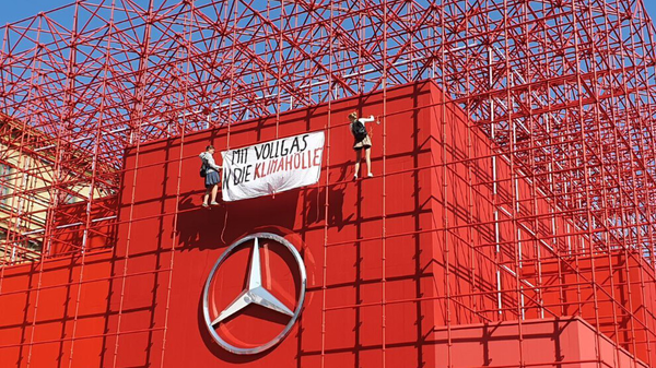 Zu sehen ist der obere Teil des Mercedes Werks in München. Gebäude ist oben mit roten Metallgittern versehen. in der Mitte zu sehen ist der Mercedes Stern im üblichen Design. Darüber zwei Kletter-Aktivistinnen mit einem Banner "Mit Vollgas die Klimahölle"