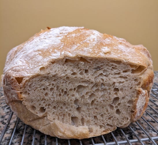 A loaf of homemade sourdough bread on a cooling rack.
The top is dusted with flour and the end is sliced off, revealing bubbles of various sizes.