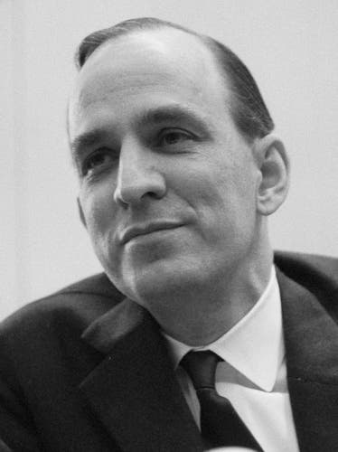 Black and white, 1966 picture. His hair is dark and straight. He is going bald. He is wearing a dark suit, a dark tie and a white shirt. 