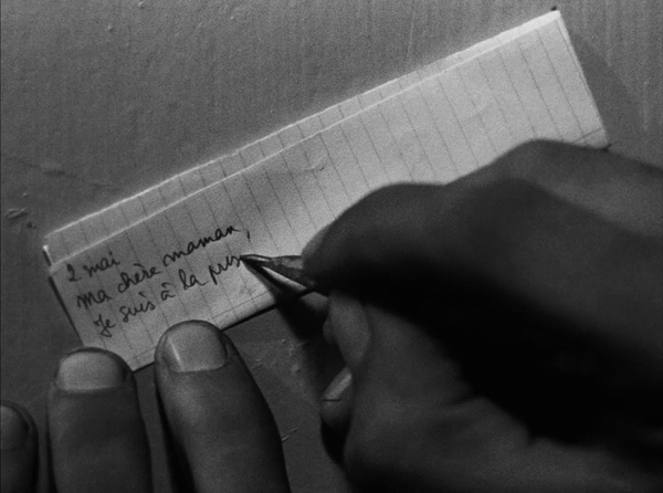 A man's hand writing a note on a scrap of paper with a short, much sharpened pencil. It starts: “2 mai  Ma chère mamam, Je suis à la pri…”