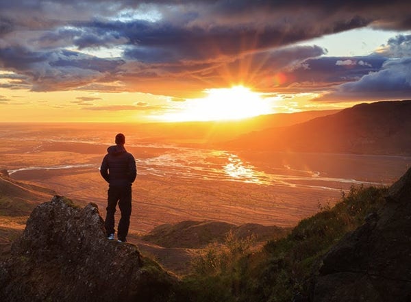 A man stands atop a peak, overlooking a beautiful sunset.