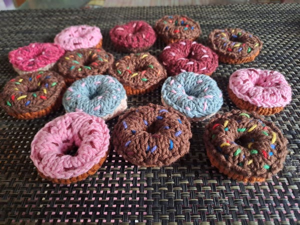 15 doughnuts, knitted, in chocolate colour with multicoloured sprinkles, and some pink and some blue doughnuts.