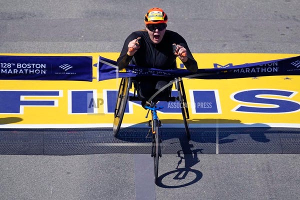 A woman in an orange bike helmet with a number bib attached, sitting in a racing wheelchair, breaks the tape at the finish line of the Boston Marathon. 