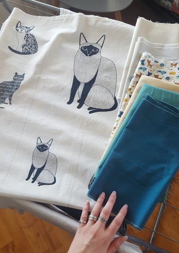 A photo of seven different fabrics laid out. One of them is a fabric with panels of black and white stylised cats on a white fabric, one of them is a white background with tiny colourful cats upon it. The rest of the fabrics are the colours of the cats in the latter mentioned: blue, teal, yellow cream, ivory, white. Sini's hand is in the frame, to provide scale and white balancing. The combination of fabrics looks like it might have potential, if correctly pondered.