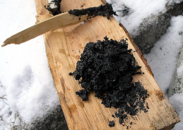 This is a photo of birch tar.

It shows a block of birch wood, cut into a plank, on which a black mass is gathered.

This is the birch tar, which has likely been burned beforehand.

The background is snowy ground.