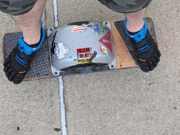 Of one wheel XR board that looks like a skateboard with a go-kart Tire in the middle and the person standing on it is wearing Vibram toe shoes with their toes hanging over the edge of the board