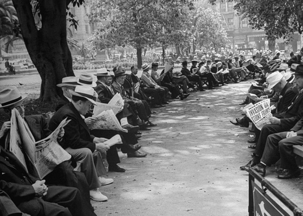 Black and white photograph on a park where two rows of men reading the newspaper. Almost all of them hav ea newspaper and are looking down to it.