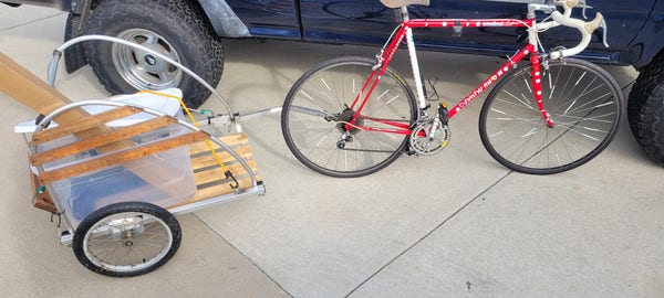 Cargo trailer attached to a red and white racing bicycle. The cargo trailer is aluminum and there is a plastic container with a shredder in it and a long box
