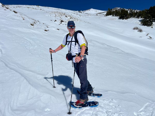 A man in a white t-shirt and blue trousers in stood in snowshoes on a white snow slope. Some trees in the upper right.