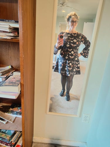 A white lady taking a mirror selfie, wear a black dress with white roses and moths. It has a boat neck, bell sleeve and ends about an inch above the knee.