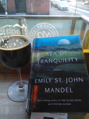 The book "Sea of Tranquility" by E. St. John Mandel on a restaurant table by a glass of porter beer.