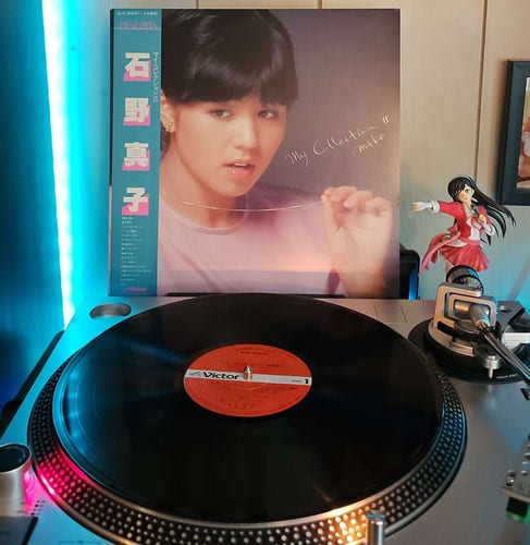 A vinyl record sits on a turntable. Behind the turntable, a vinyl album outer sleeve is displayed. The front cover shows Mako Ishino holding her necklace out in front of her as she looks down and away from the camera.. 