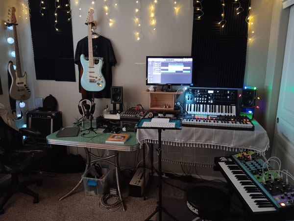 Fender Jazz bass, a couple Peavey guitar amps, (closed) laptop, hand-assembled custom Stratocaster, Sennheiser HD6XX headphones, Shure 57 microphone, 3" Mackie monitors, Behringer X1204 mixer, Yamaha THR30II guitar amp, Mac Mini (with trackpad and keyboard on a movable stand), a MIDIRock "Stone" joystick (unfortunately obscured by the mac peripherals), Korg Wavestate, Akai MPK249 controller keyboard, Moog Matriarch, Peace hydraulic drum throne.
