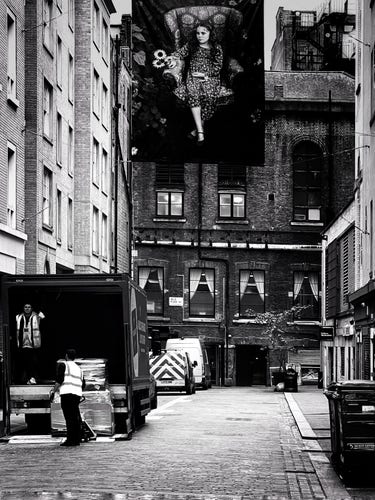 A black and white photo taken in the so-called Soho Photography Quarter in Ramillies Street in London's Soho district. Looking down the street, on all three sides are the back sides of buildings situated on surrounding streets. On the left are two guys delivering goods from a truck. High above the middle of the street, hanging from a cable, is an 8-foot by 4-foot full-height portrait of a girl of about 10 years of age in a flowery dress, sitting in a floral-patterned armchair, holding three sunflowers and surrounded by foliage. She is looking at the building that is receiving the delivery with a somewhat bored expression
