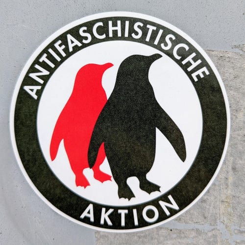 Ein runderAufkleber "Antifaschistische Aktion". Innerhalb eines schwarzen Kreises befinden sich ein roter und ein schwarzer Pinguin