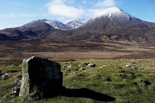 A small upright block of stone standing on top of a grassy mound in the foreground. The backdrop is a group of three mountain summits, grey-green at the lower slopes but snow-capped. It's very bright and sunny.