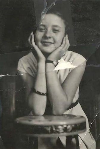 A young girl standing behind a stool. Her elbows are resting on the chair and she is holding her head in her hands.